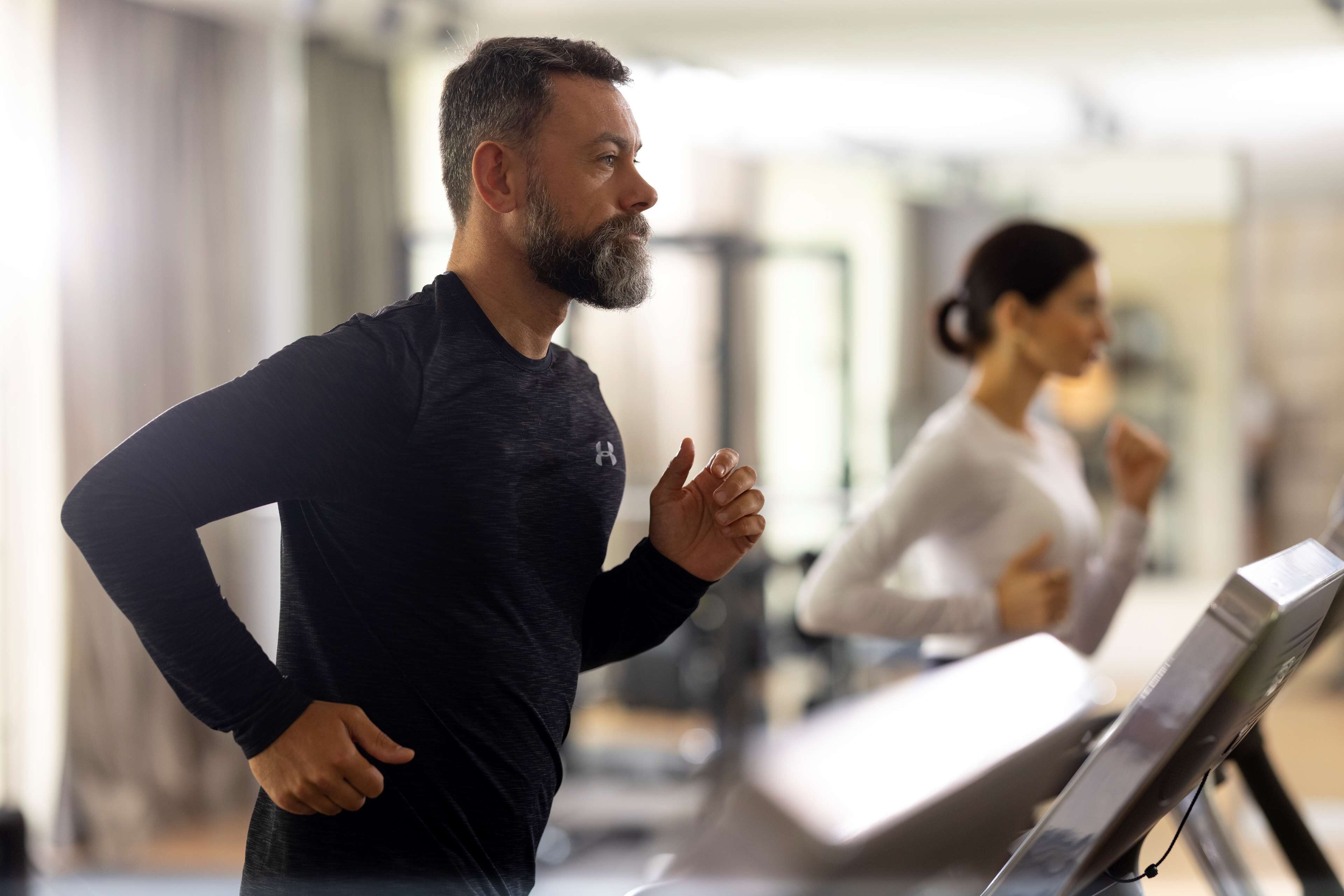 A man in a black long-sleeve shirt running on a treadmill in a gym with a woman in the background.