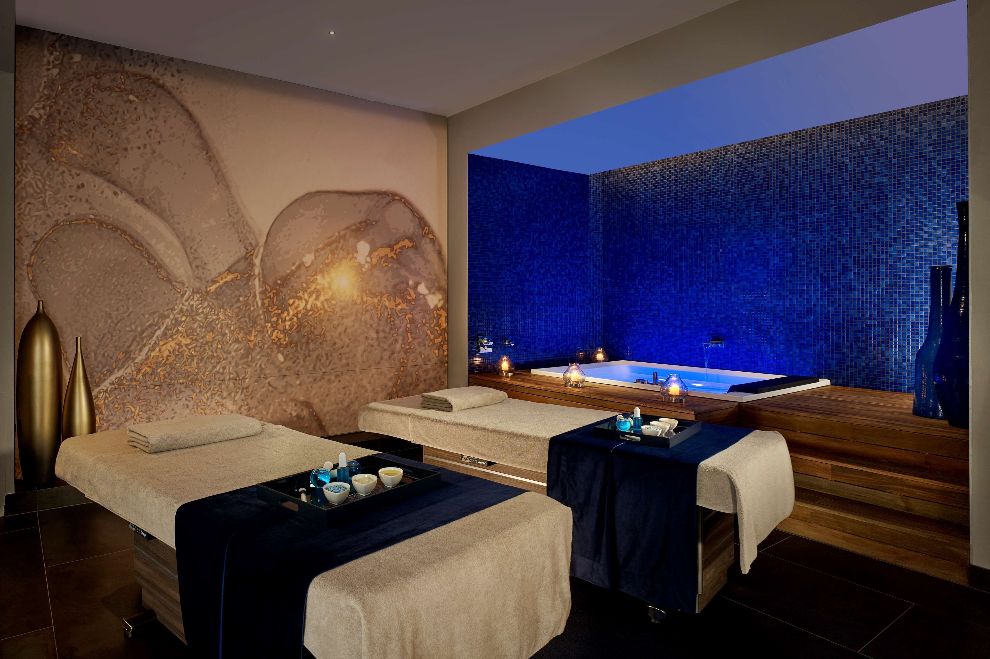 Une salle de spa équipée de deux tables de massage, d’une grande fresque murale, d’un mur décoré de mosaïques bleues et d’un bain à remous.
