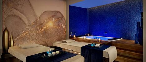 Une salle de spa équipée de deux tables de massage, d’une grande fresque murale, d’un mur décoré de mosaïques bleues et d’un bain à remous.