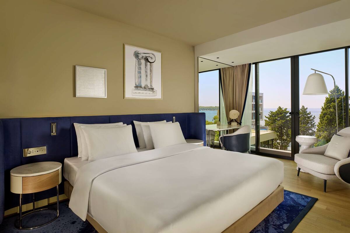 A modern hotel room with a large bed, a desk, and a chair. There are two framed pictures on the wall, and a view of the sea through large windows.