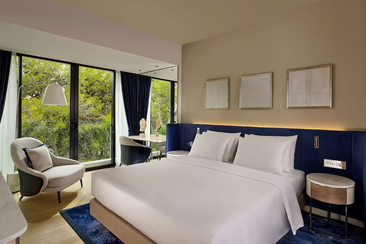 A modern hotel room with a large bed, a desk, a chair, and a view of greenery through large windows.