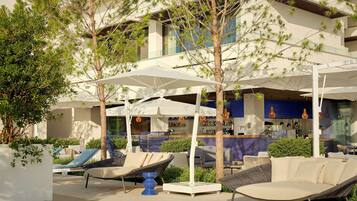 Un área de estar al aire libre con sillones reclinables y sombrillas, un bar con decoración azul y blanca, y un edificio de varios pisos con grandes ventanales.