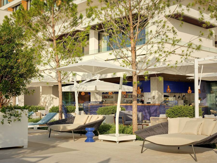 Un área de estar al aire libre con sillones reclinables y sombrillas, un bar con decoración azul y blanca, y un edificio de varios pisos con grandes ventanales.