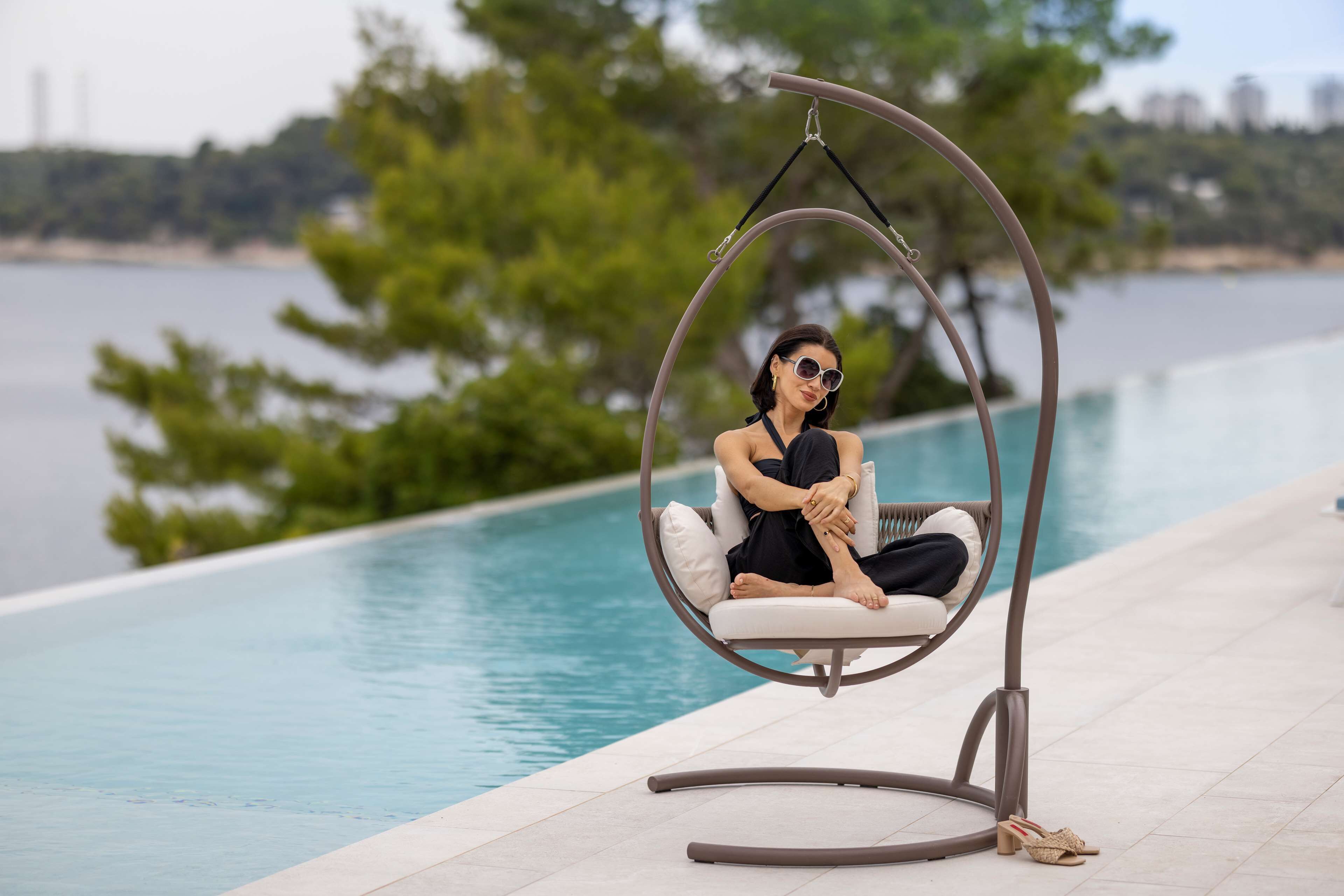 A person sitting in a hanging chair by a pool with a view of the water and trees.