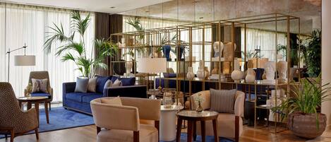 A modern living room with a blue sofa, beige armchairs, and a coffee table. There is a large mirror on the wall and several potted plants.