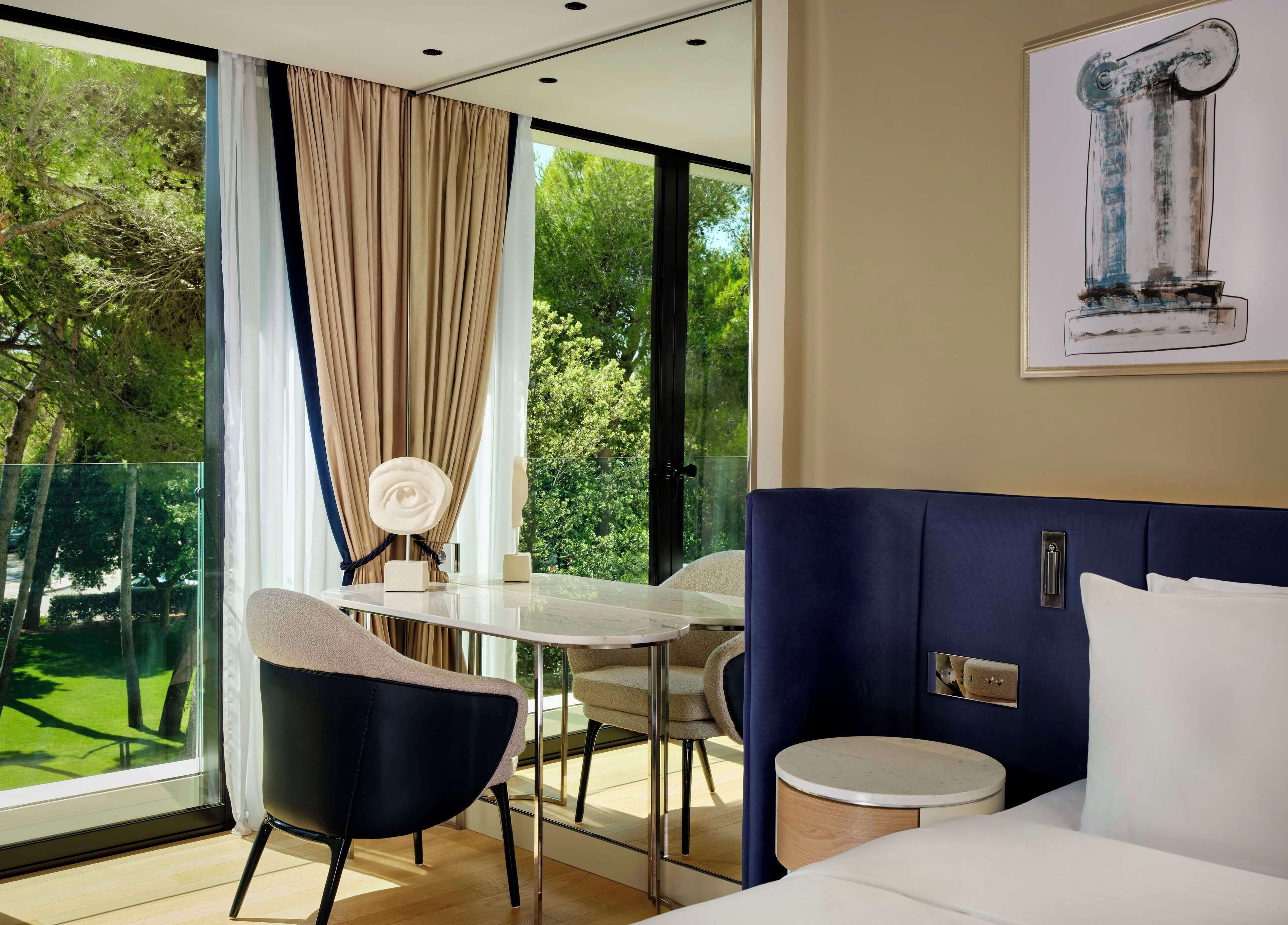 A modern hotel room with a large window, a dining table, two chairs, a bed, and a painting of a column.
