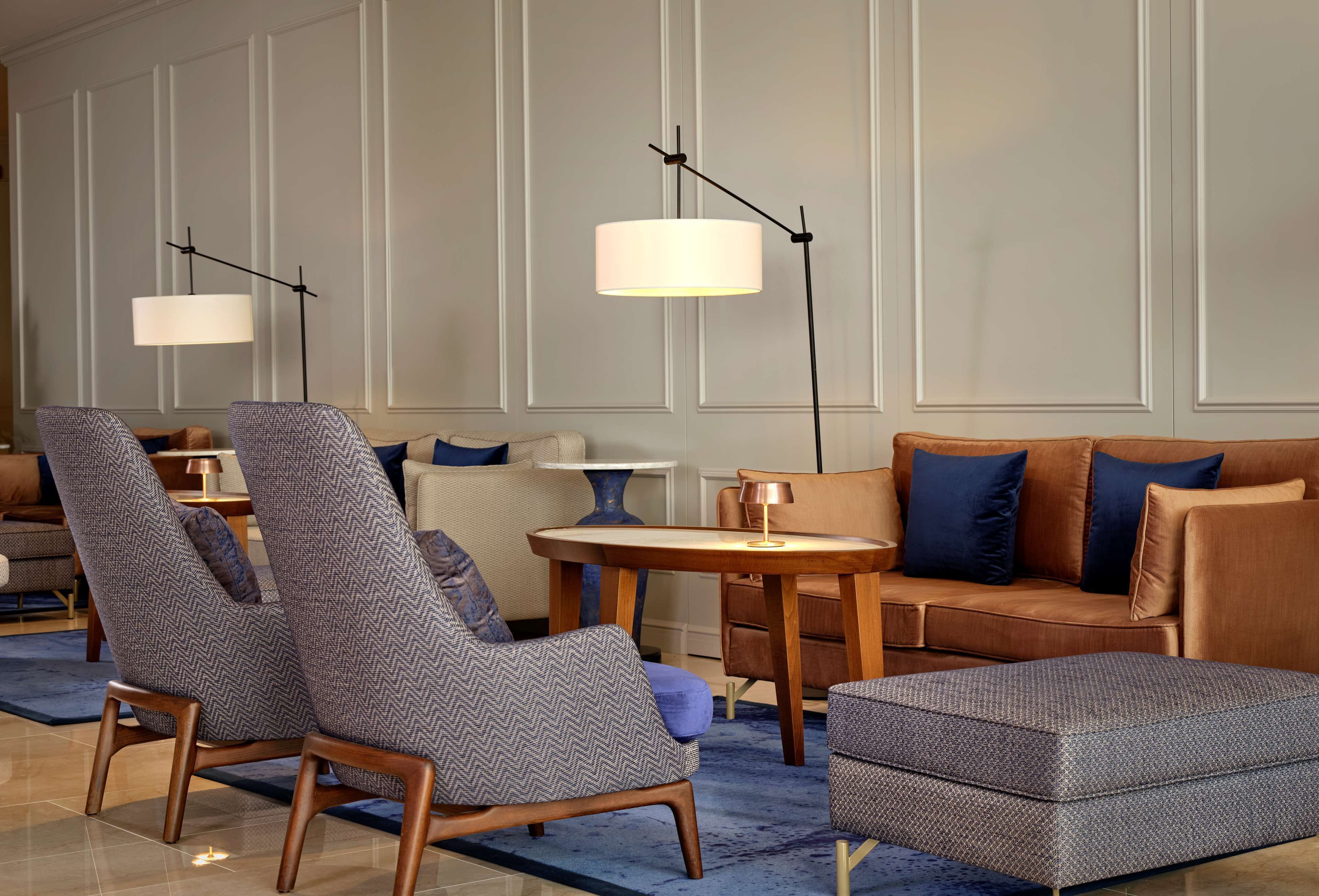 A modern hotel lobby with a seating area featuring patterned armchairs, a brown sofa, and a round table with a lamp.