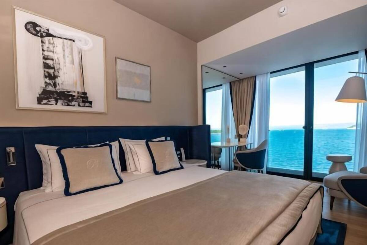 A hotel room with a large bed, a view of the sea, a dining area, and a balcony.