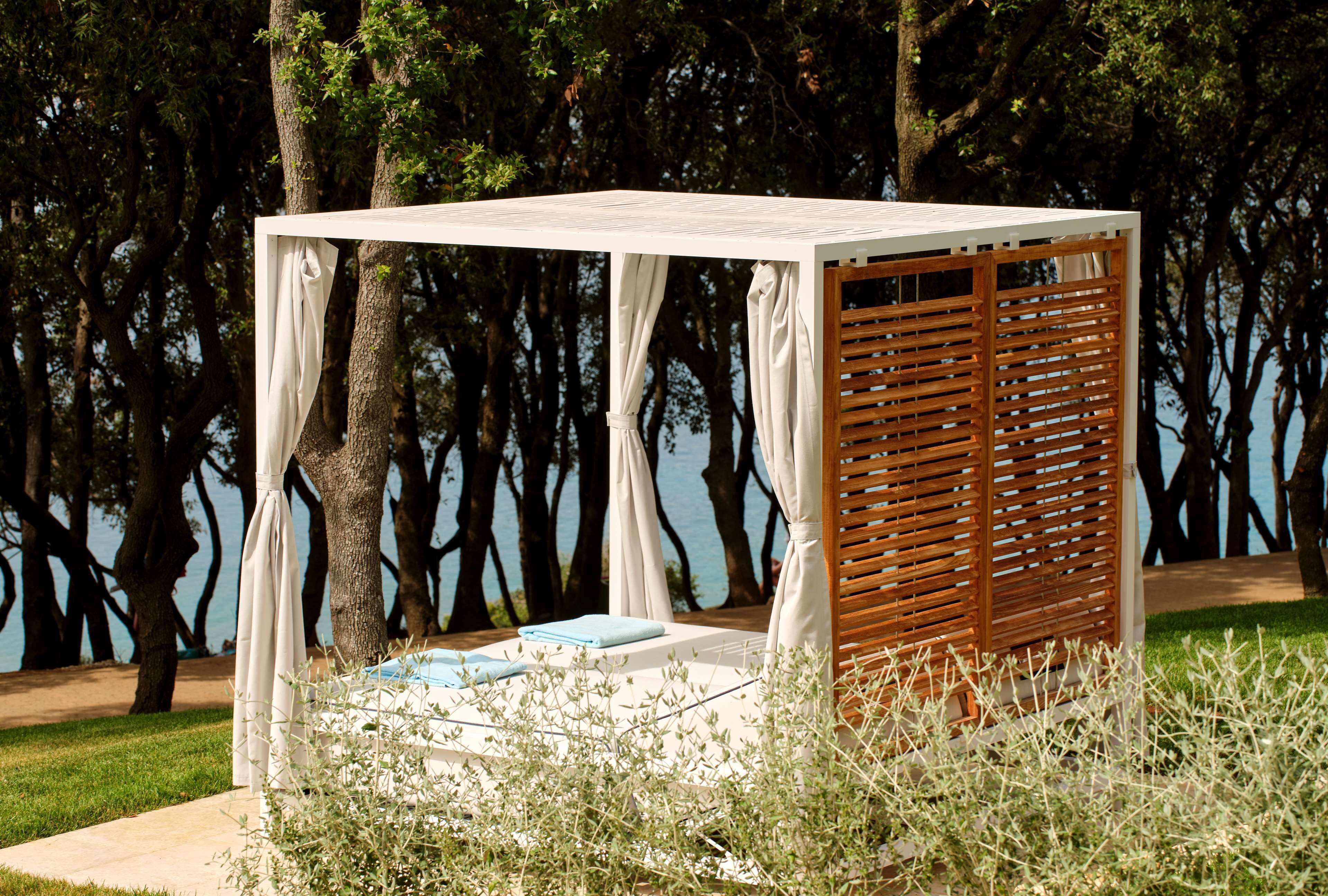 A covered outdoor seating area with a wooden slatted wall, a cushioned bench, and a small table with a blue towel.