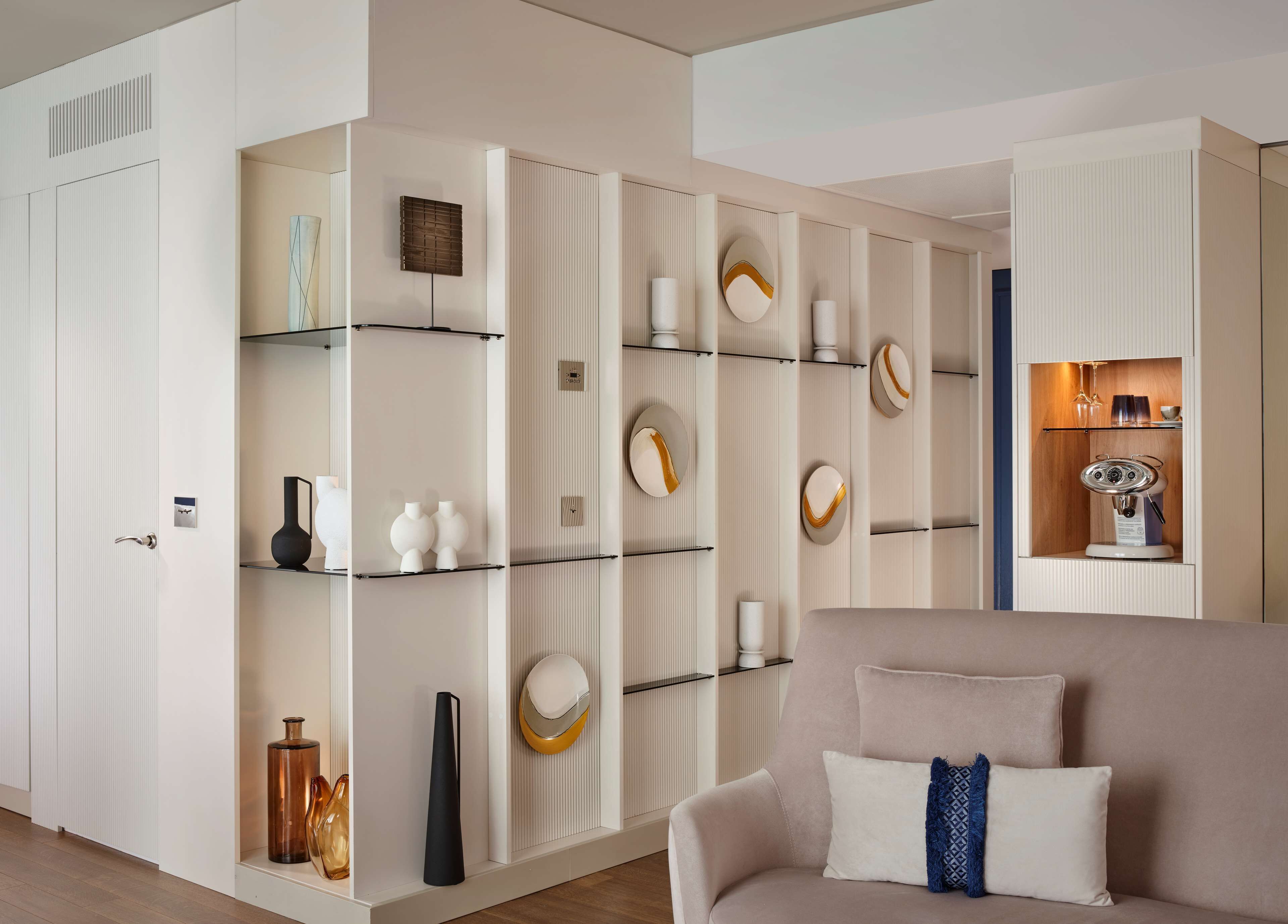 A modern living room with a sofa, a coffee machine, and a shelving unit with decorative items.
