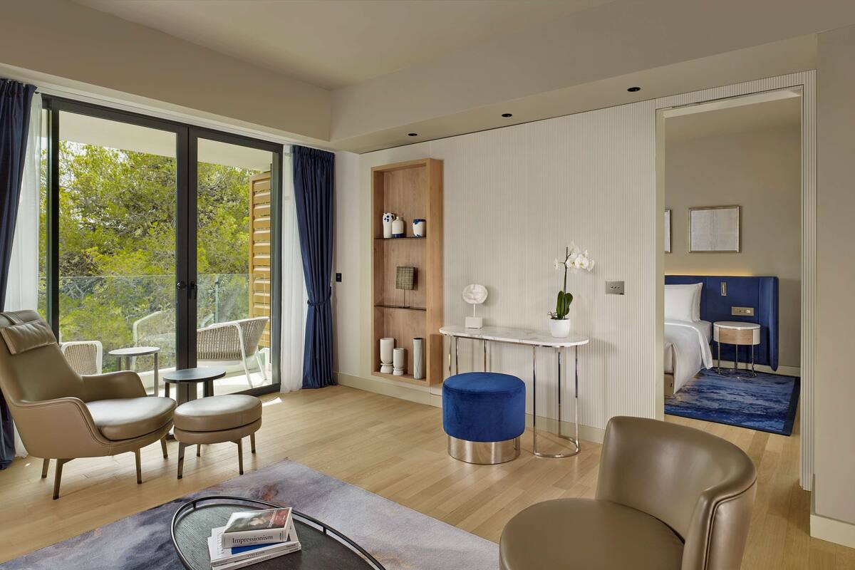 A modern hotel room with a balcony, a seating area with a sofa and chairs, a small table, and a bed with a headboard.
