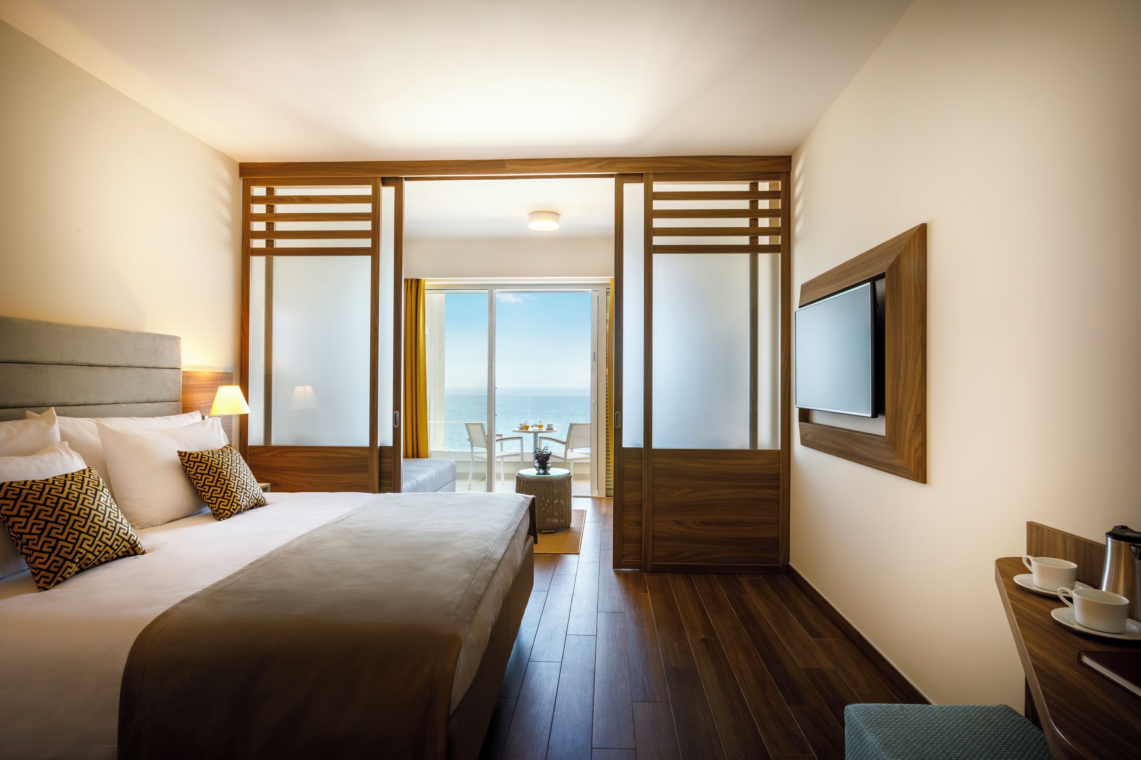 A hotel room with a bed, a TV, a desk with a coffee maker, and a view of the sea.