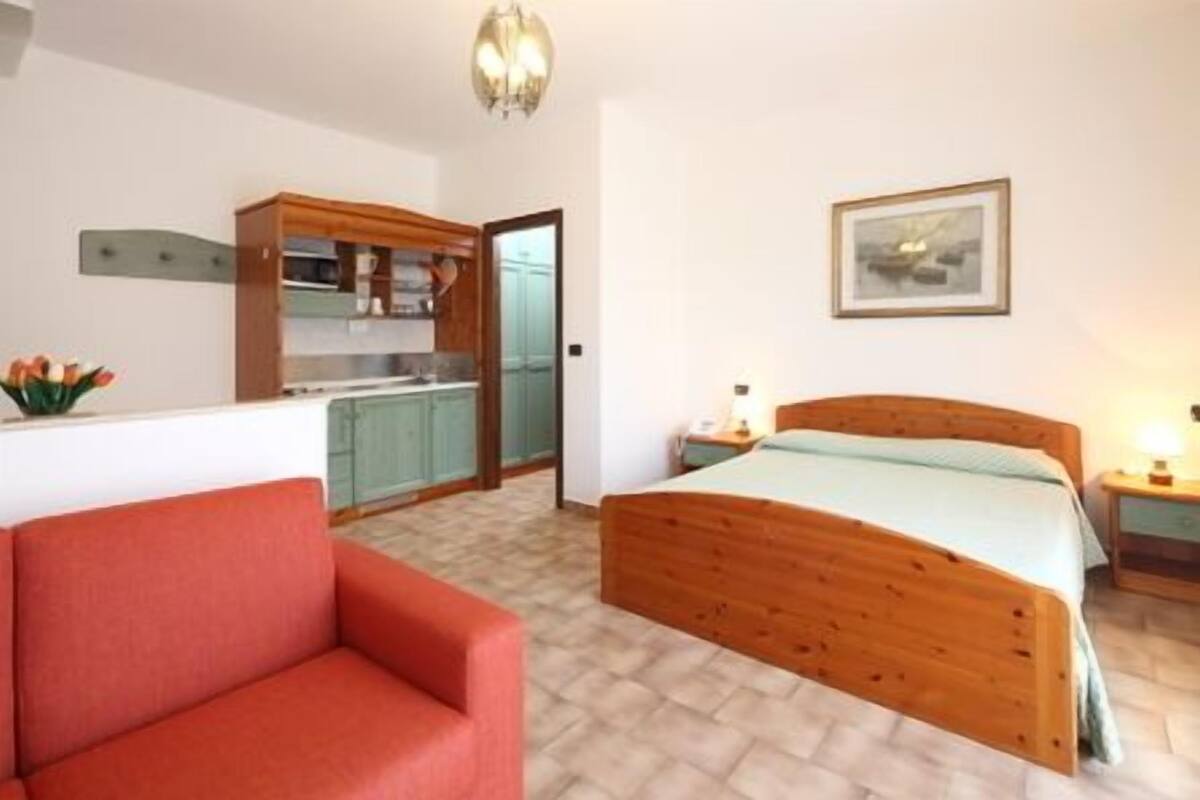 A bedroom with a wooden bed, a red sofa, a kitchenette, and a painting on the wall.