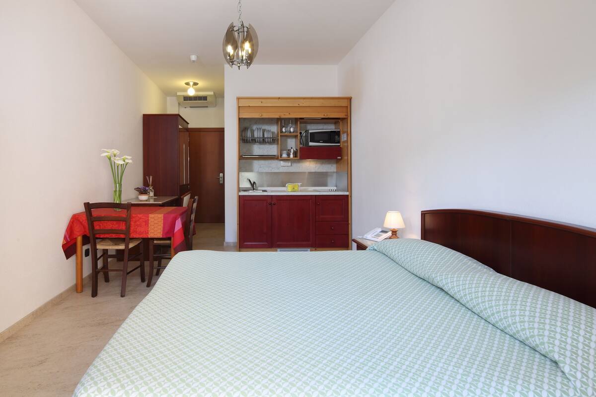 A bedroom with a large bed, a wooden headboard, a nightstand with a lamp, and a small kitchenette with a microwave and cabinets.
