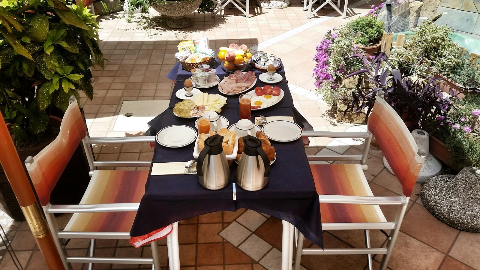 Mesa lista para el desayuno con diversos platos, entre ellos huevos, pan y bebidas, en un patio exterior con sillas y plantas en macetas.