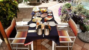 Ein Frühstückstisch mit verschiedenen Speisen, darunter Eier, Brot und Getränke, auf einer Terrasses mit Stühlen und Topfpflanzen.