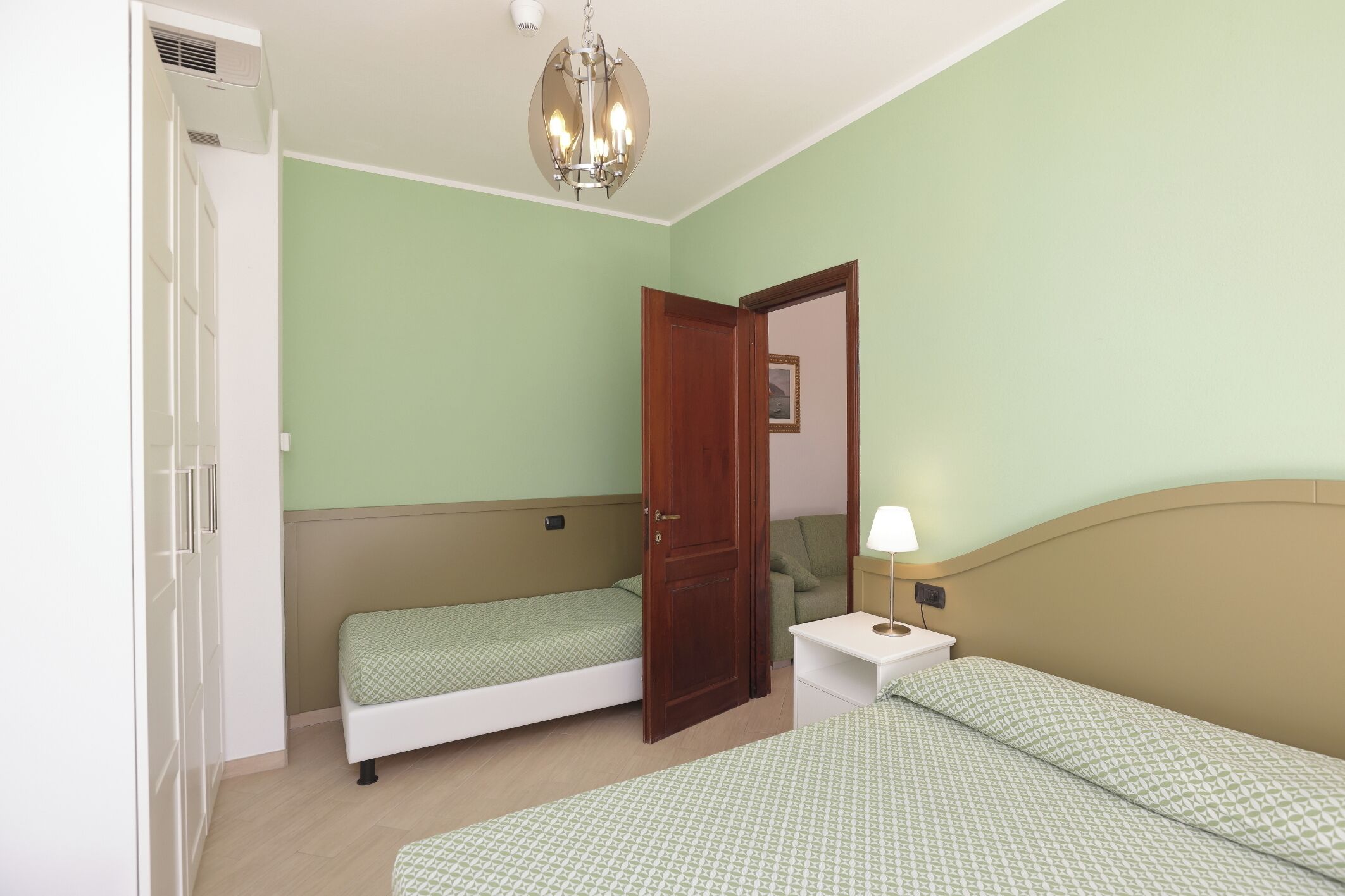 Quarto com duas camas, um guarda-roupa, mesa de cabeceira com luminária e ventilador de teto.