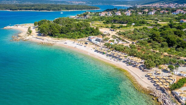 An aerial view of a coastal area with a beach, a marina, and a residential area with houses. - Hotel Kanajt (Punat)