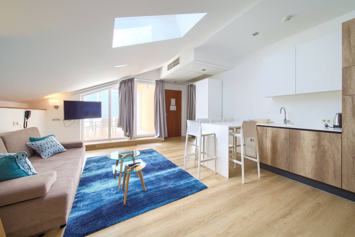 Una sala de estar moderna con un sofá, una mesa de centro y un televisor. Se observa una zona de cocina con una barra y una ventana con cortinas.