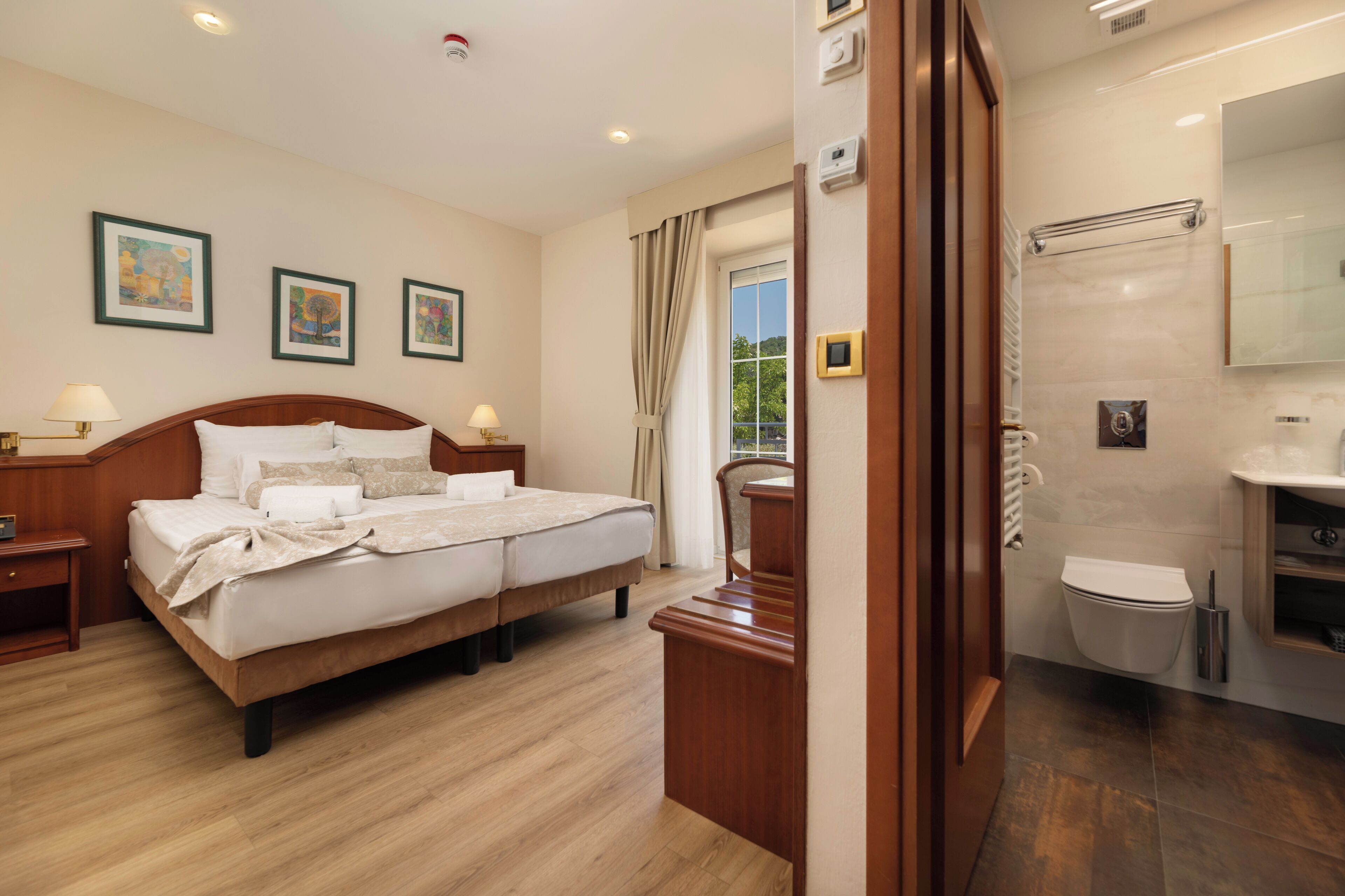 A hotel room with a wooden bed, a nightstand, a bedside table, a bathroom with a toilet, a sink, and a mirror.
