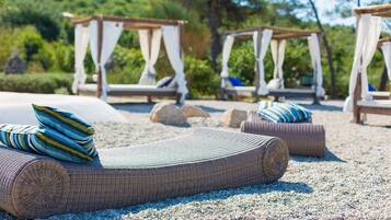 A beachside lounge area with wicker loungers, pillows, and a pebbled ground.