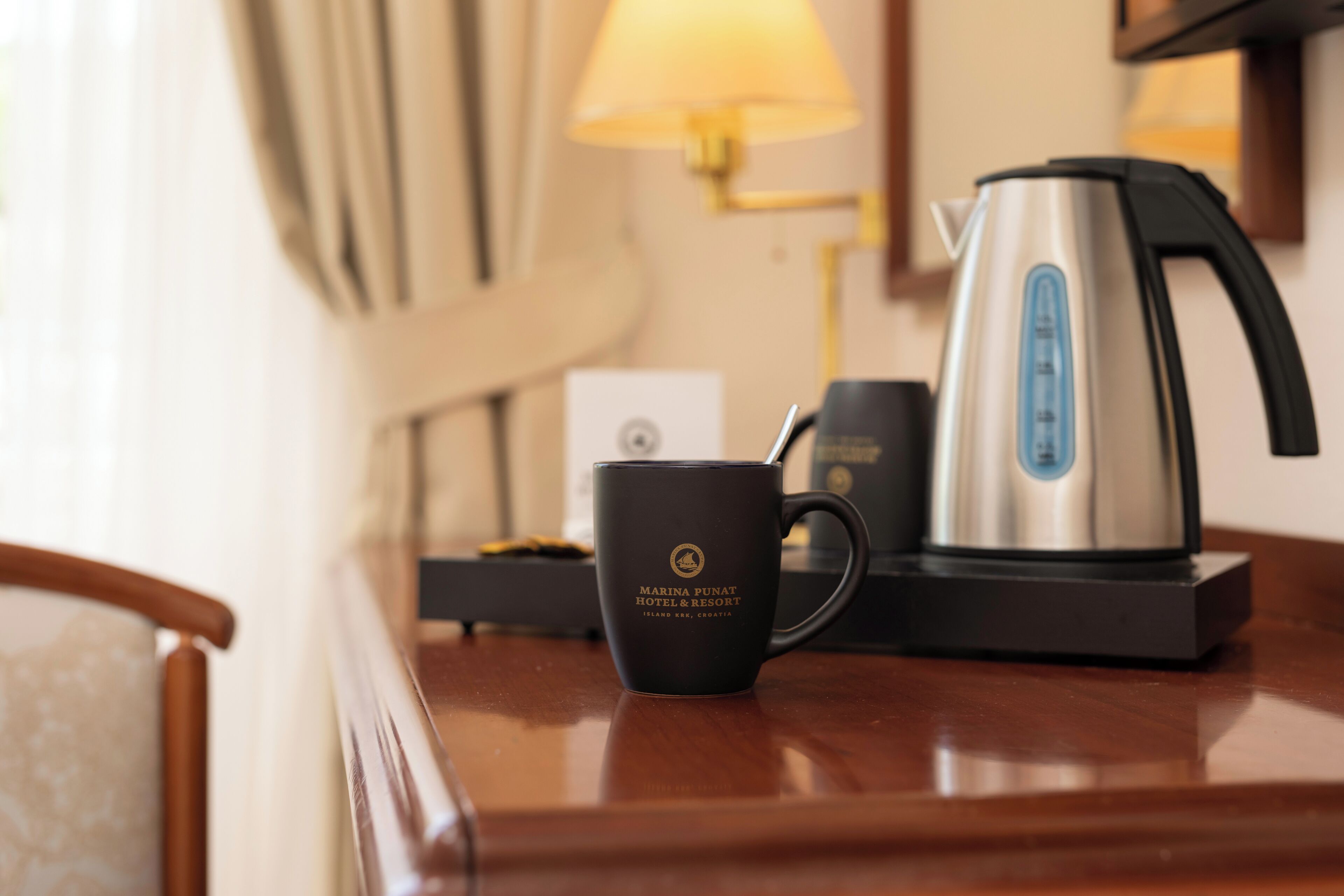 Una cafetera, una taza y una lámpara sobre una mesa de madera en una habitación de hotel.