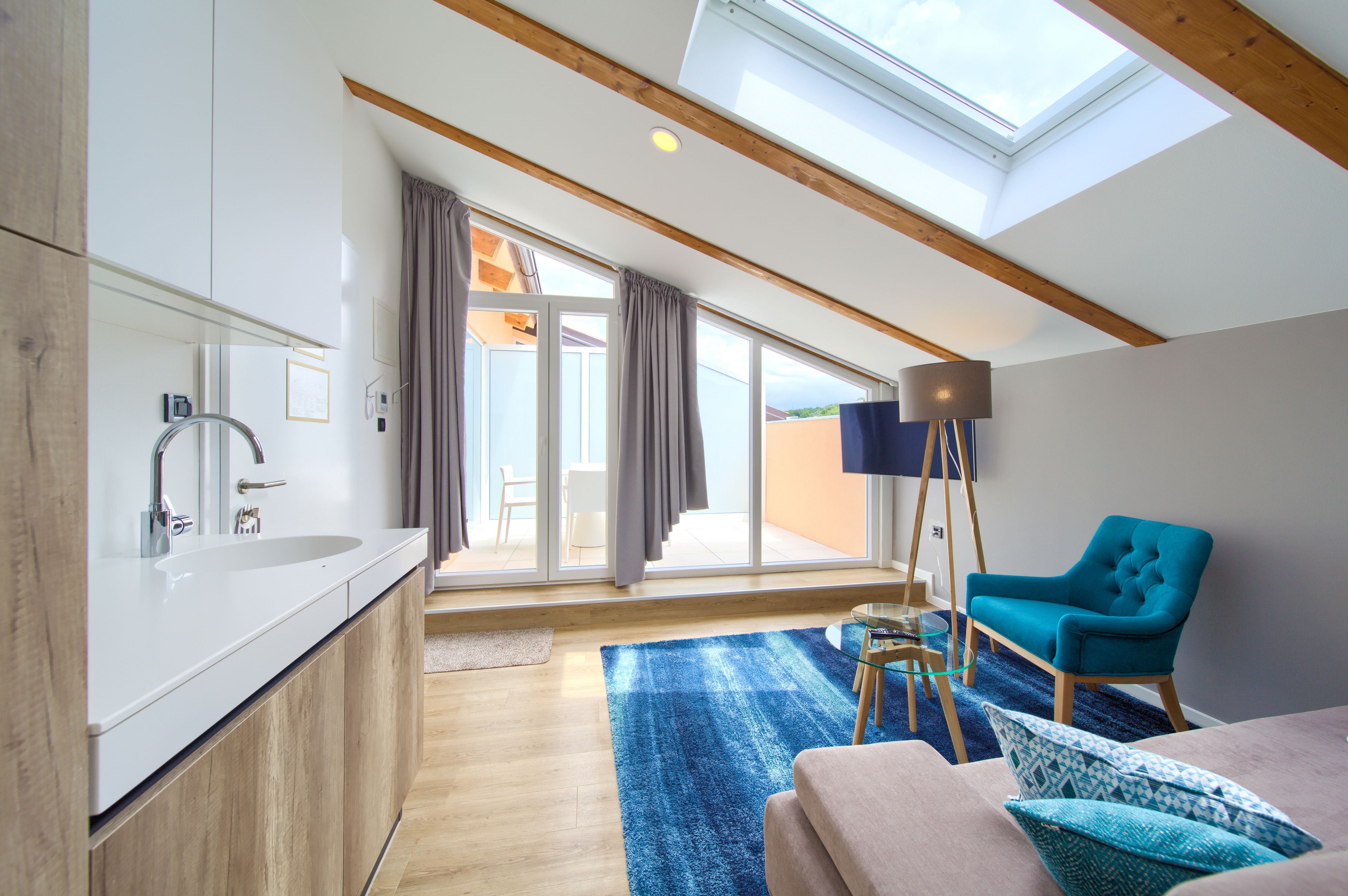 A modern kitchen with a skylight, a large window with curtains, a blue rug, and a teal armchair.
