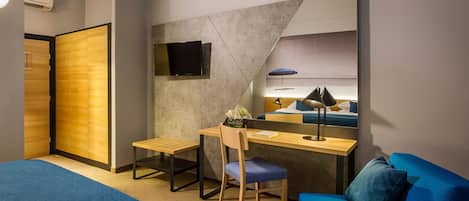 A modern hotel room with a wooden wardrobe, a desk with a chair, a blue sofa, a large mirror, and a TV mounted on the wall.