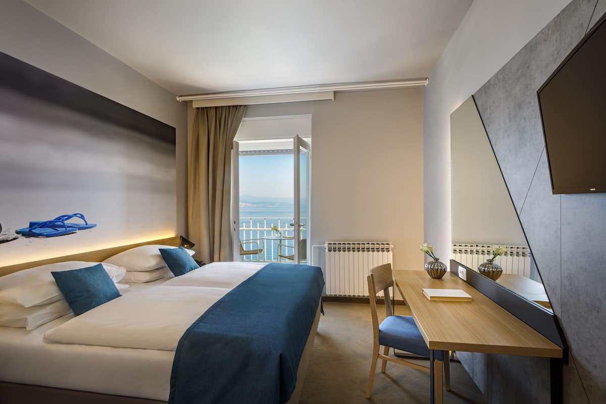 A hotel room with two beds, a wooden desk, a chair, a large mirror, and a view of the sea through an open door.
