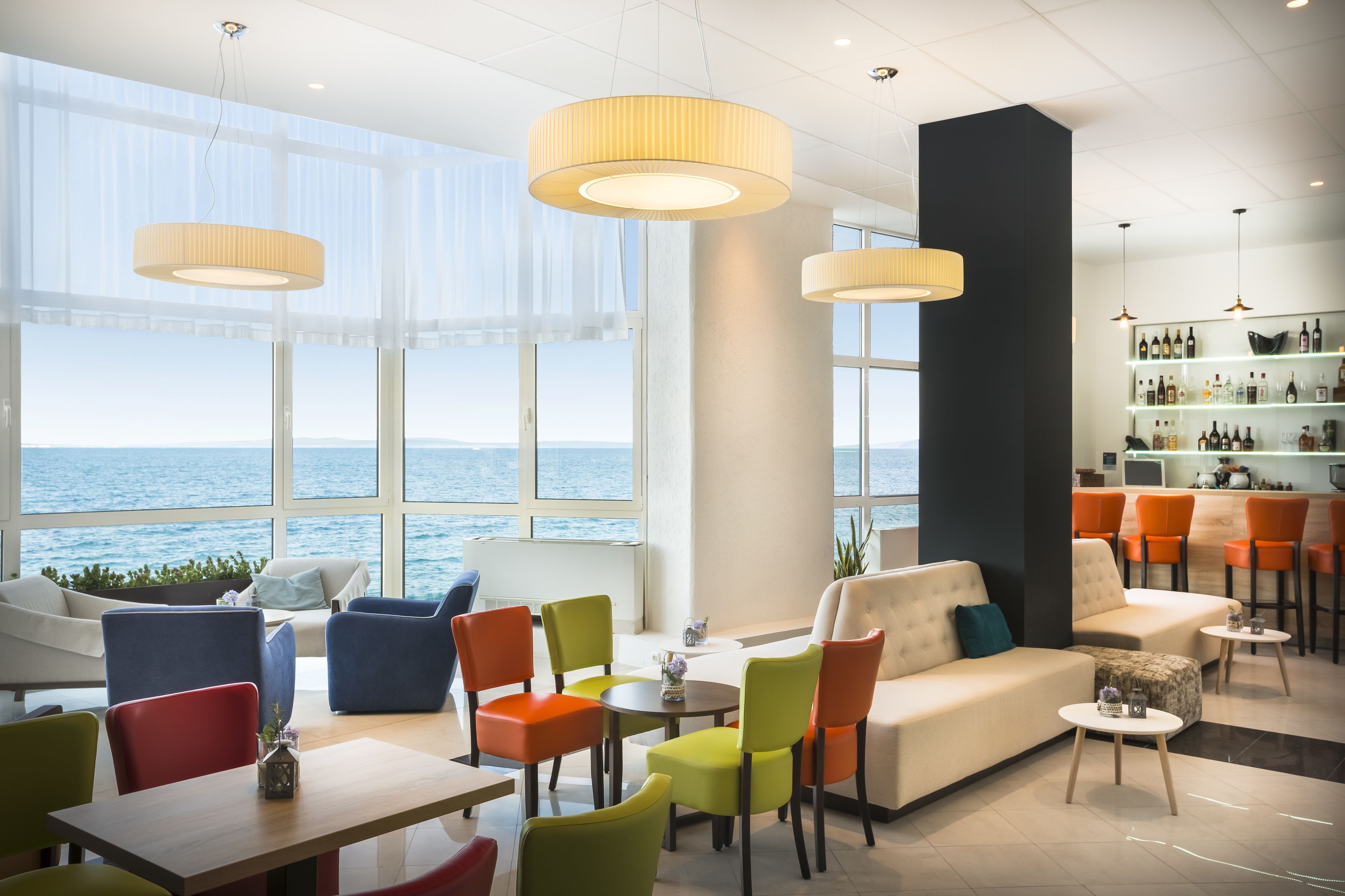 Un salon moderne avec vue sur la mer, comprenant des fauteuils colorés, un canapé et un bar avec des étagères remplies de bouteilles.
