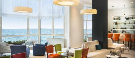 Un salon moderne avec vue sur la mer, comprenant des fauteuils colorés, un canapé et un bar avec des étagères remplies de bouteilles.