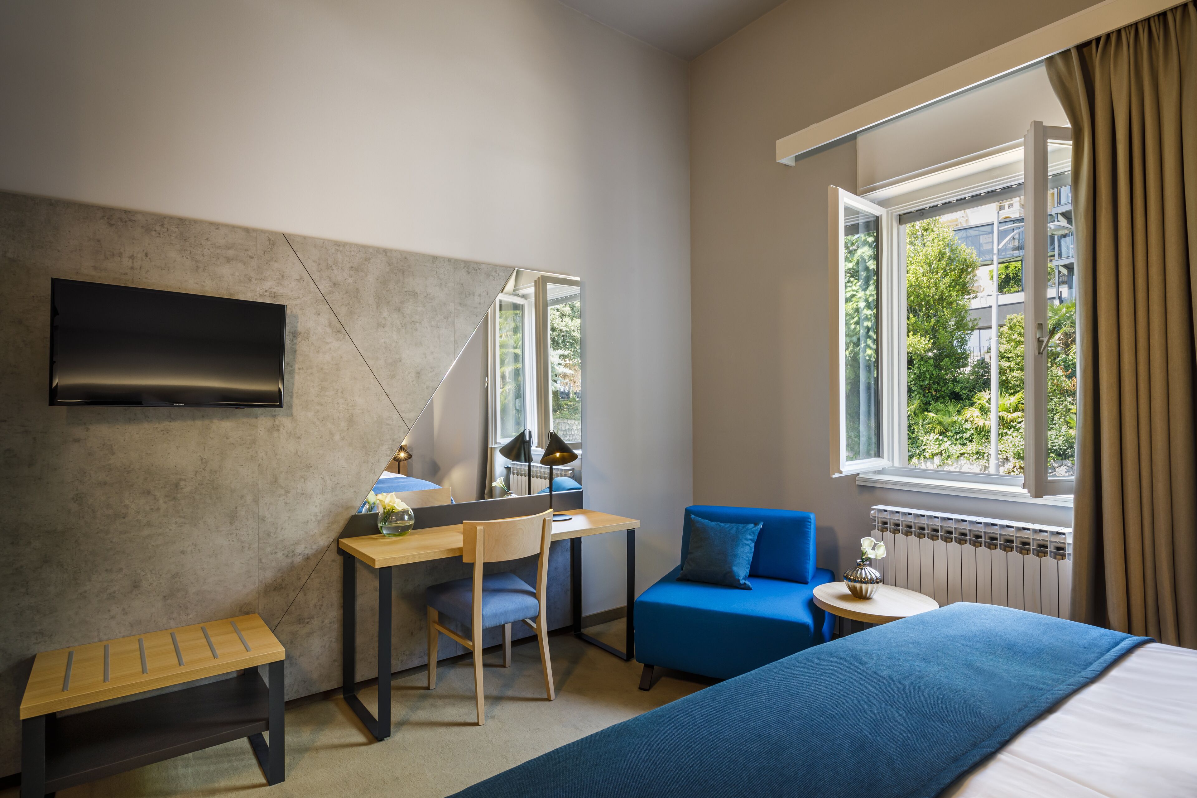 Hotellihuone, jossa on sänky, työpöytä tuolilla, pieni pöytä maljakolla ja ikkuna, jossa on verhot.