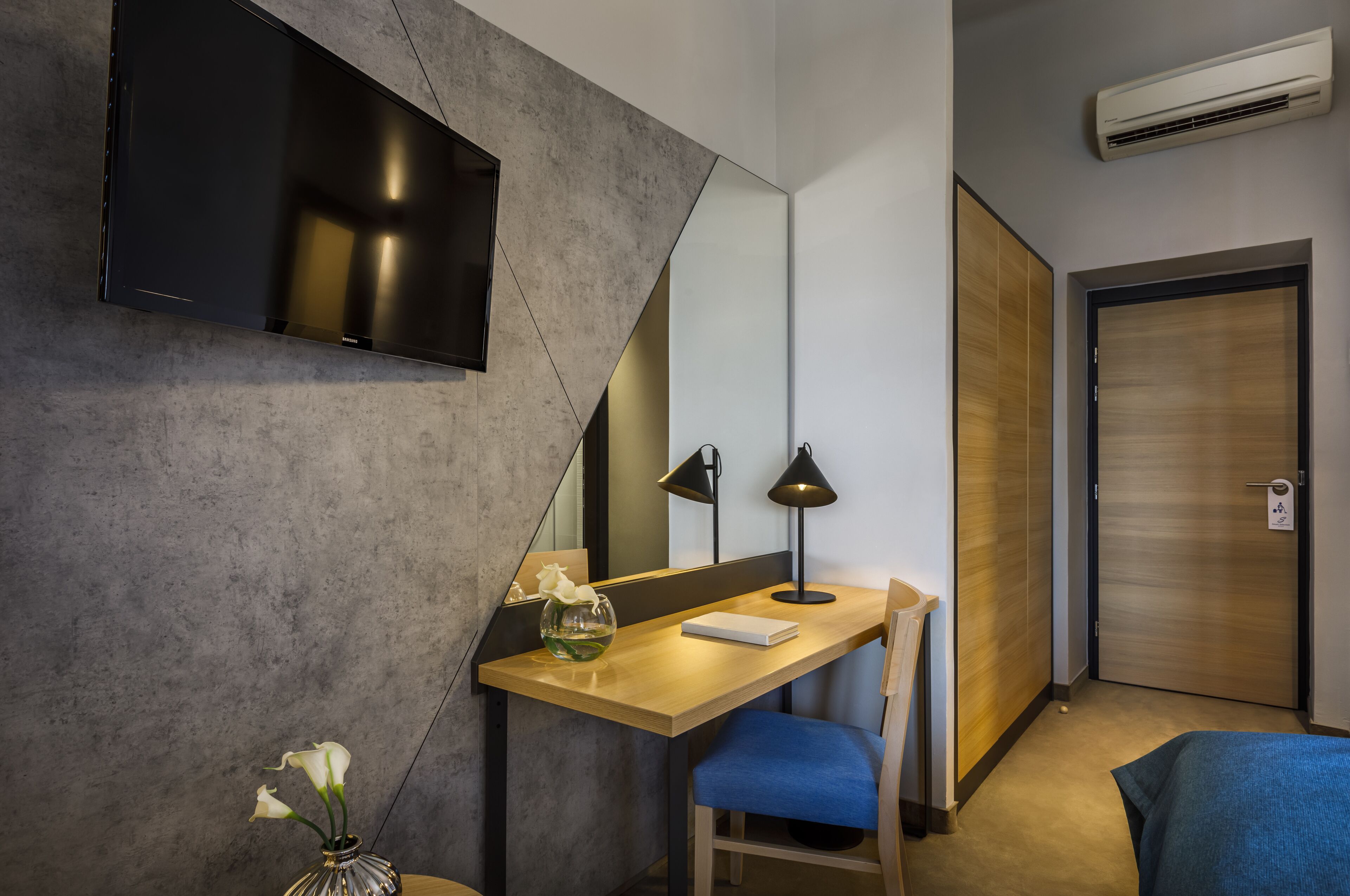 Une chambre d’hôtel moderne équipée d’un téléviseur à écran plat fixé au mur, d’un bureau avec une lampe, d’une chaise et d’un grand miroir.