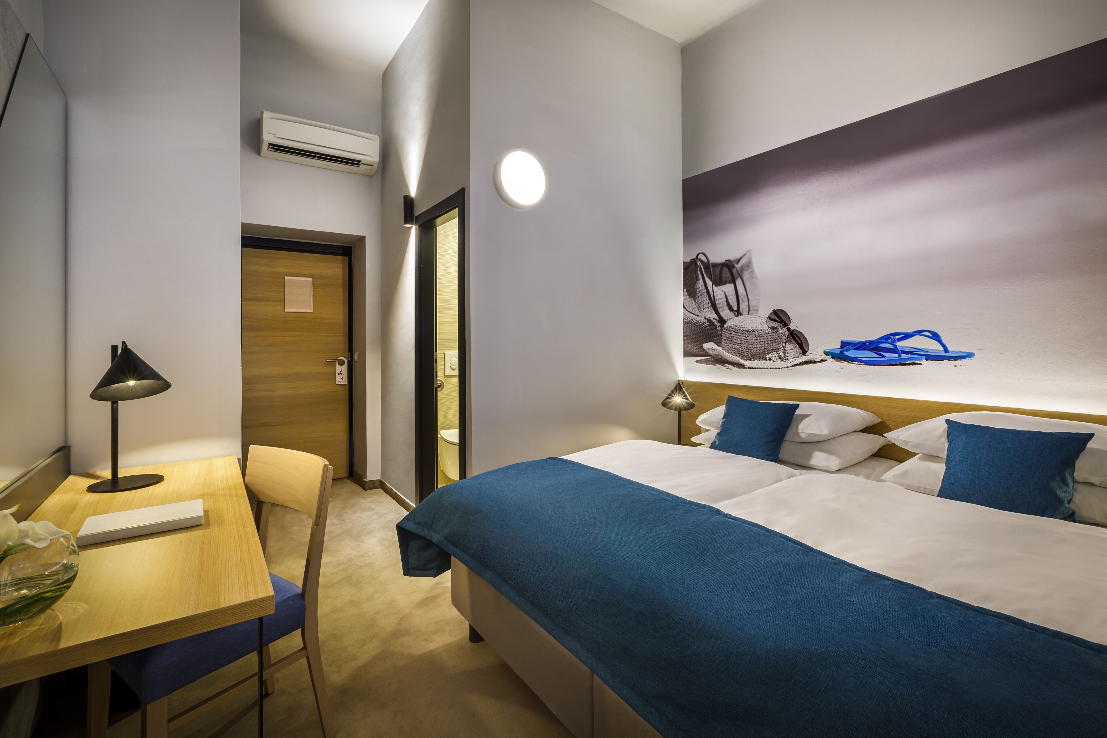 A hotel room with a bed, a desk, a chair, a lamp, and a wall-mounted shelf with a handbag and flip-flops.
