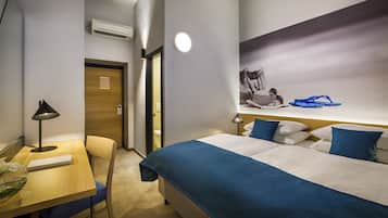 Una habitación de hotel con cama, escritorio, silla, lámpara y estante de pared con un bolso y chanclas.