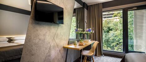 A modern hotel room with a bed, a desk with a chair, a flat-screen TV mounted on a wall, and a large window with curtains.