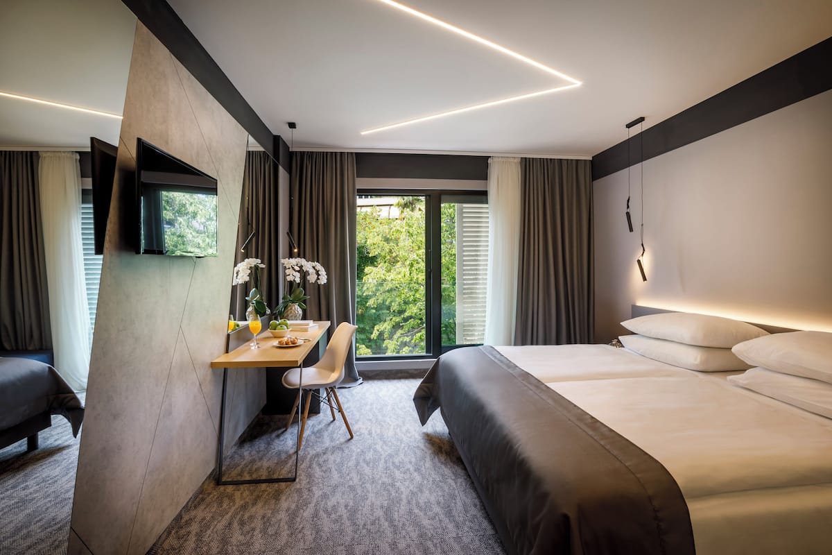 A modern hotel room with a large bed, a desk with a chair, a flat-screen TV mounted on the wall, and a large window offering a view of greenery outside.
