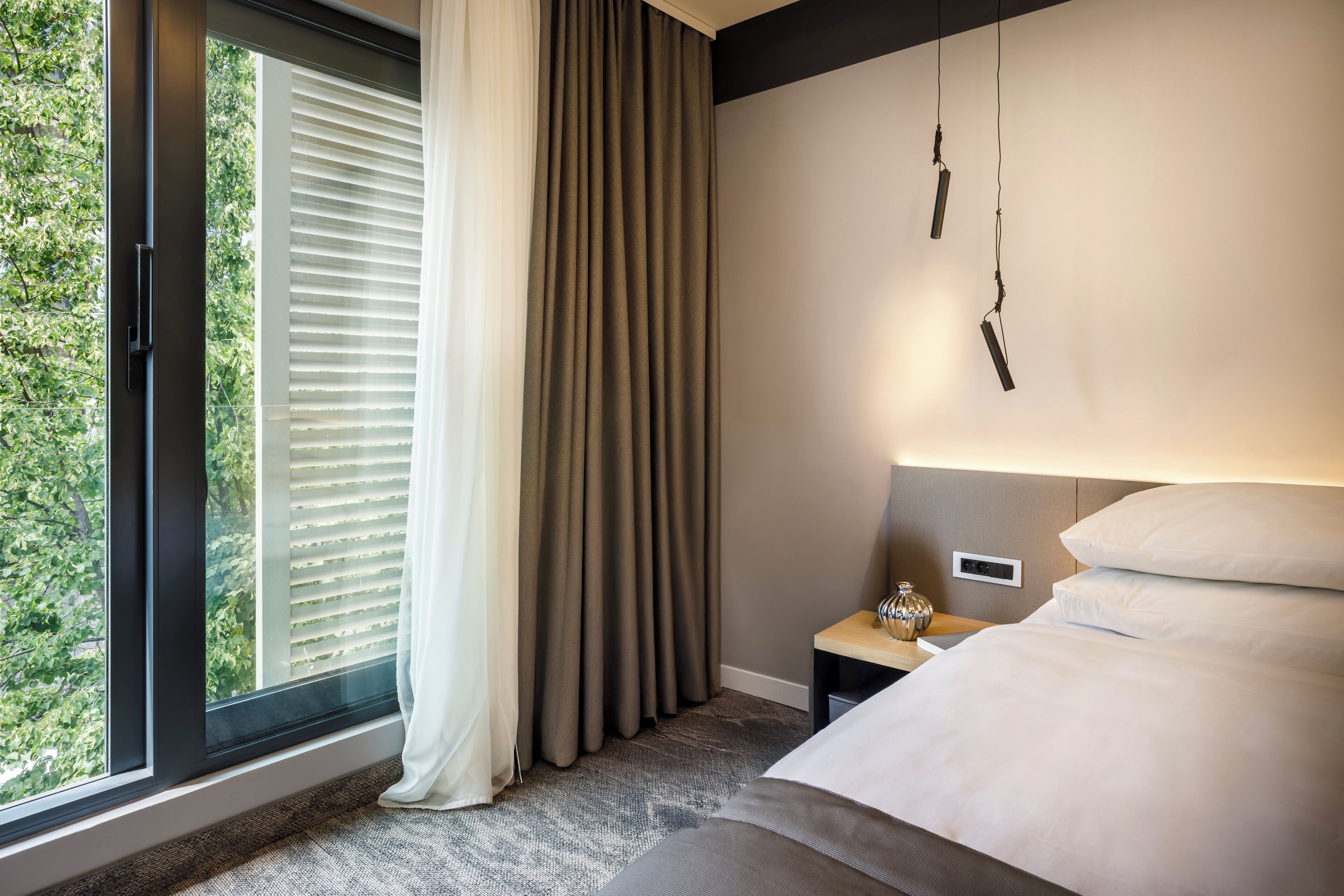 Una habitación de hotel moderna con un ventanal grande, una cama, una mesita de noche y una lámpara fijada a la pared.