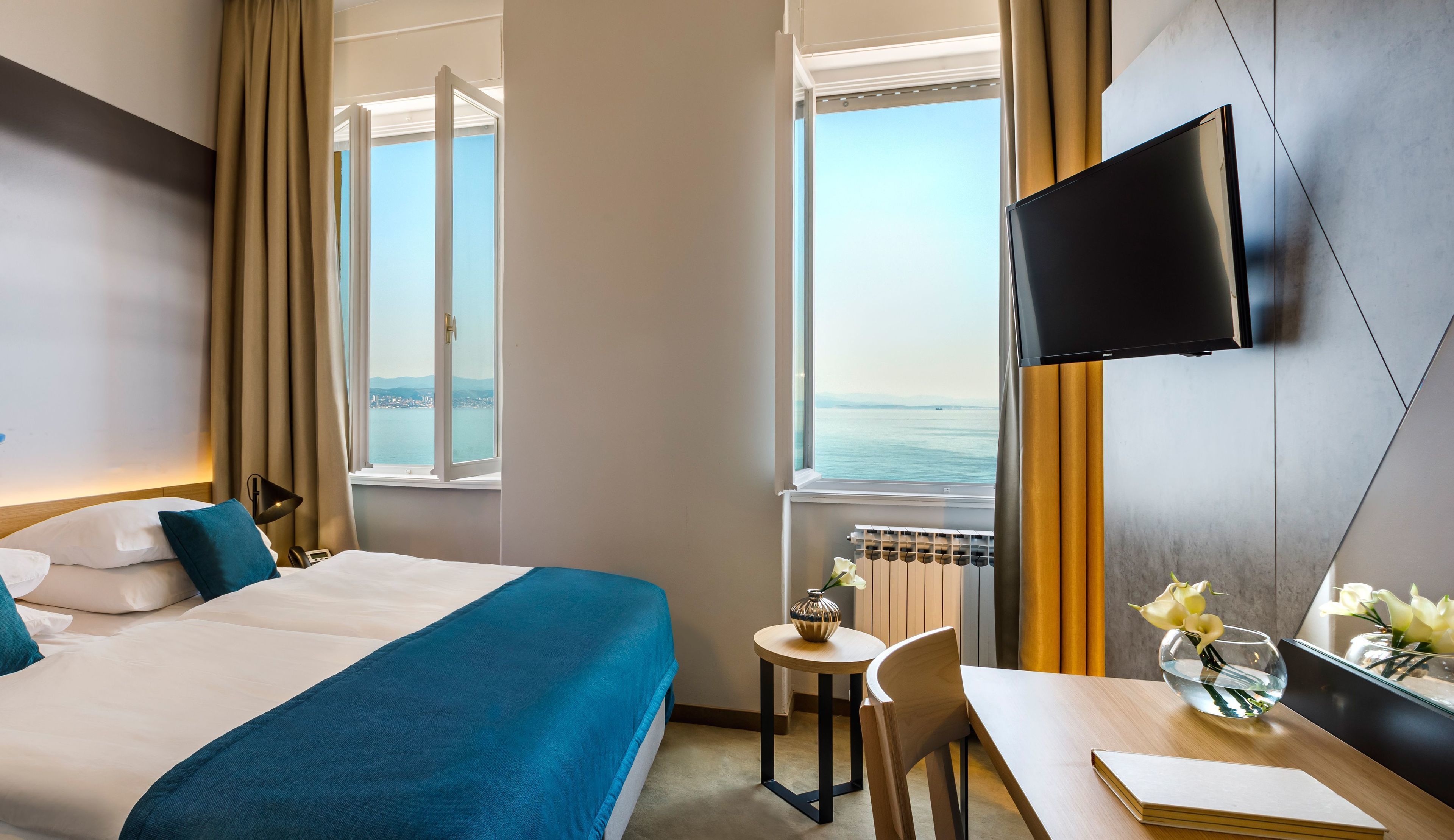 A hotel room with a bed, a desk, a chair, a TV, and a window with a view of the sea.