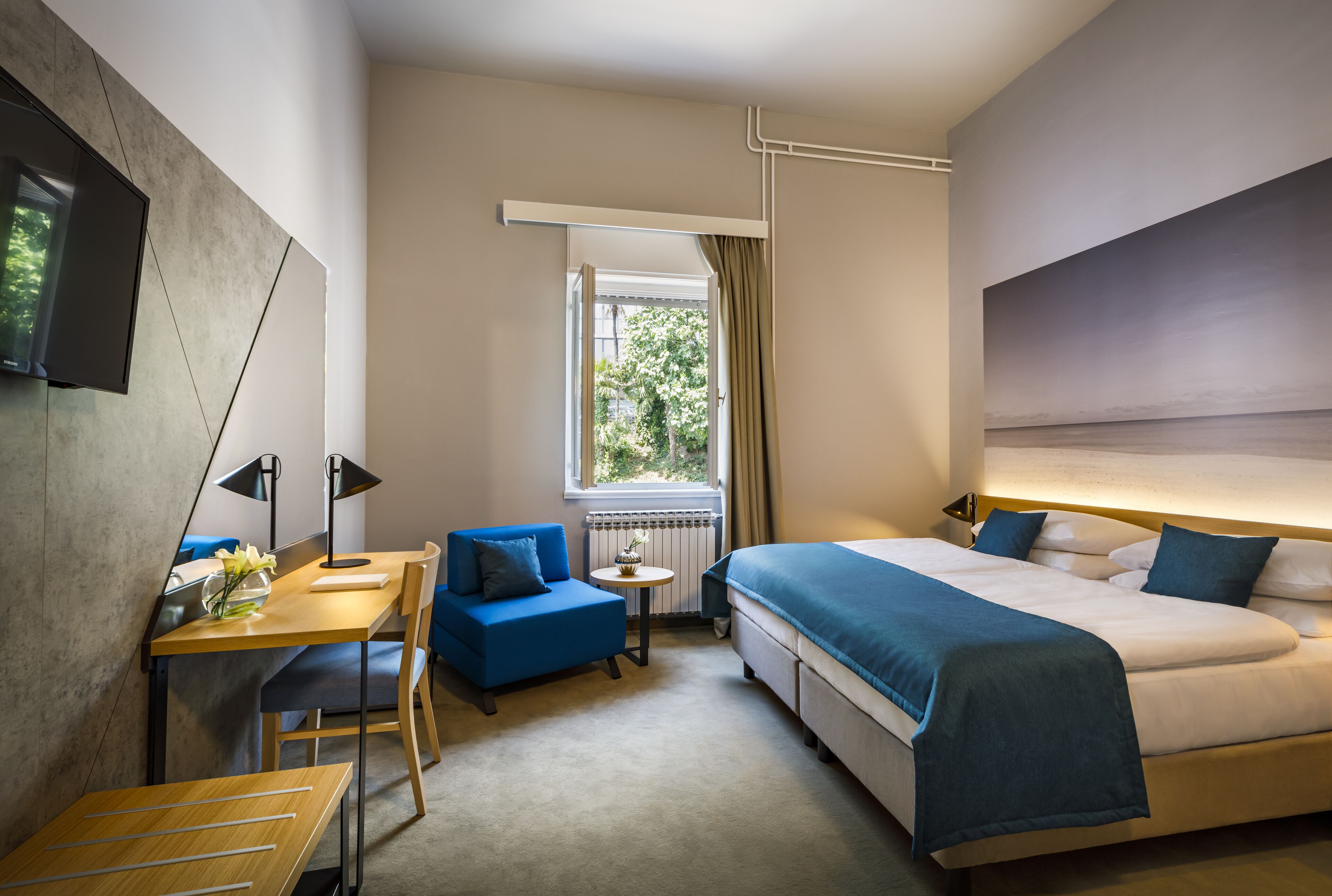 Habitación de hotel con cama, escritorio, silla, mesita y ventana con cortinas.