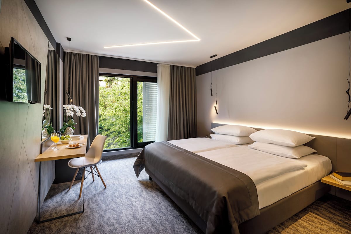 A modern hotel room with a large bed, a desk with a chair, a small table with a fruit bowl, and a view of greenery outside.