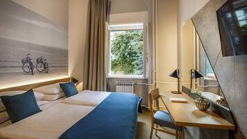 Habitación de hotel con cama, escritorio, silla, ventana con vista a árboles y espejo montado en la pared.