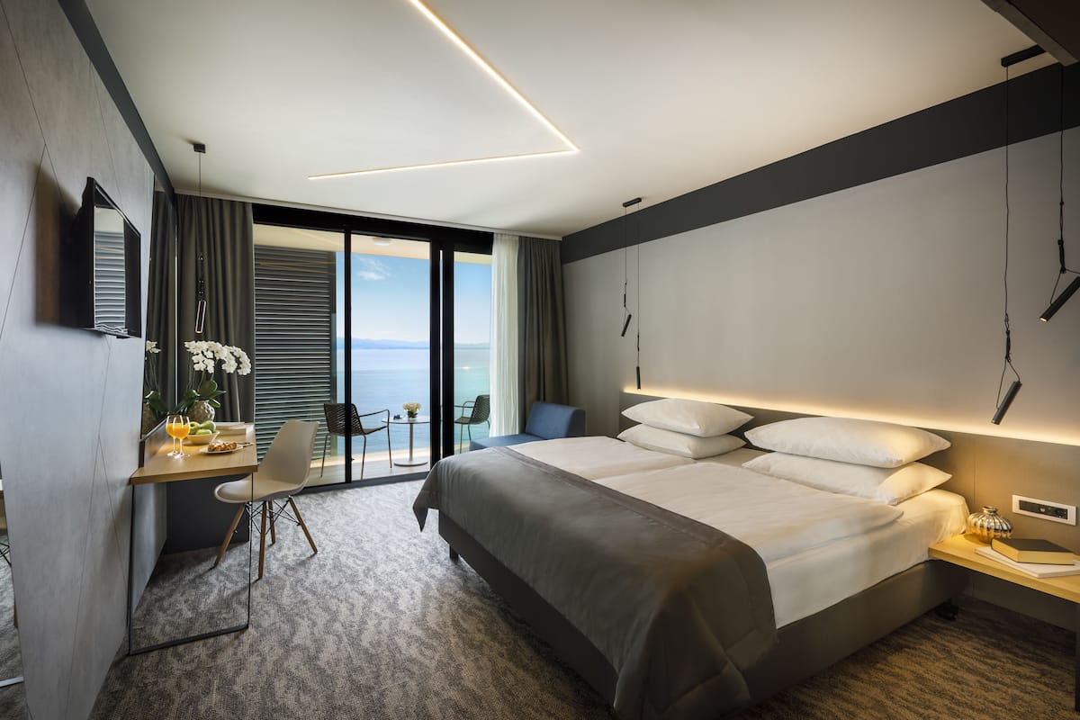 A modern hotel room with a large bed, a desk with a chair, a small table with a plant, and a view of the sea through large windows.