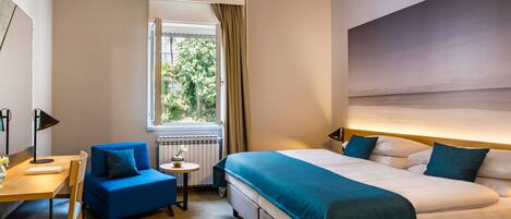 Quarto de hotel com uma cama grande, uma escrivaninha com cadeira, uma mesinha e uma janela com cortinas.