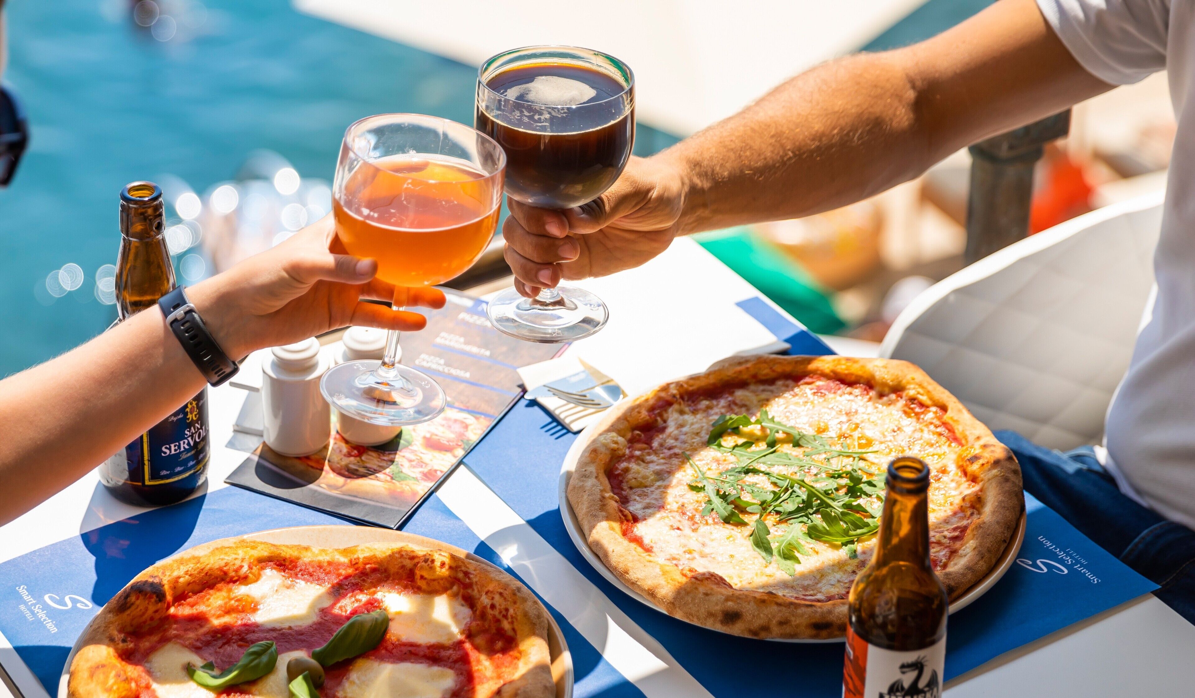 Una mesa con dos pizzas, una botella y vasos de bebidas.