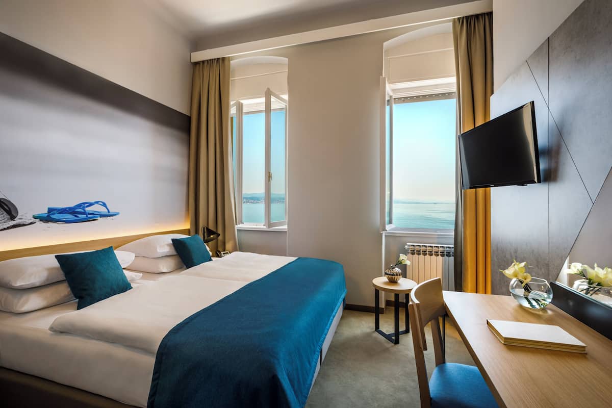 A hotel room with a bed, a desk, a chair, a TV, and a view of the sea.
