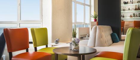 A modern lounge area with colorful chairs and a sofa, a round table, and a view of the sea through large windows.