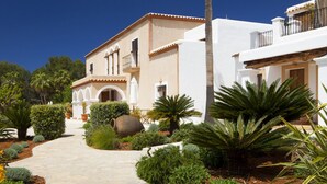 Una villa de estilo mediterráneo con un camino empedrado que lleva a la entrada, rodeada de exuberante vegetación y palmeras.