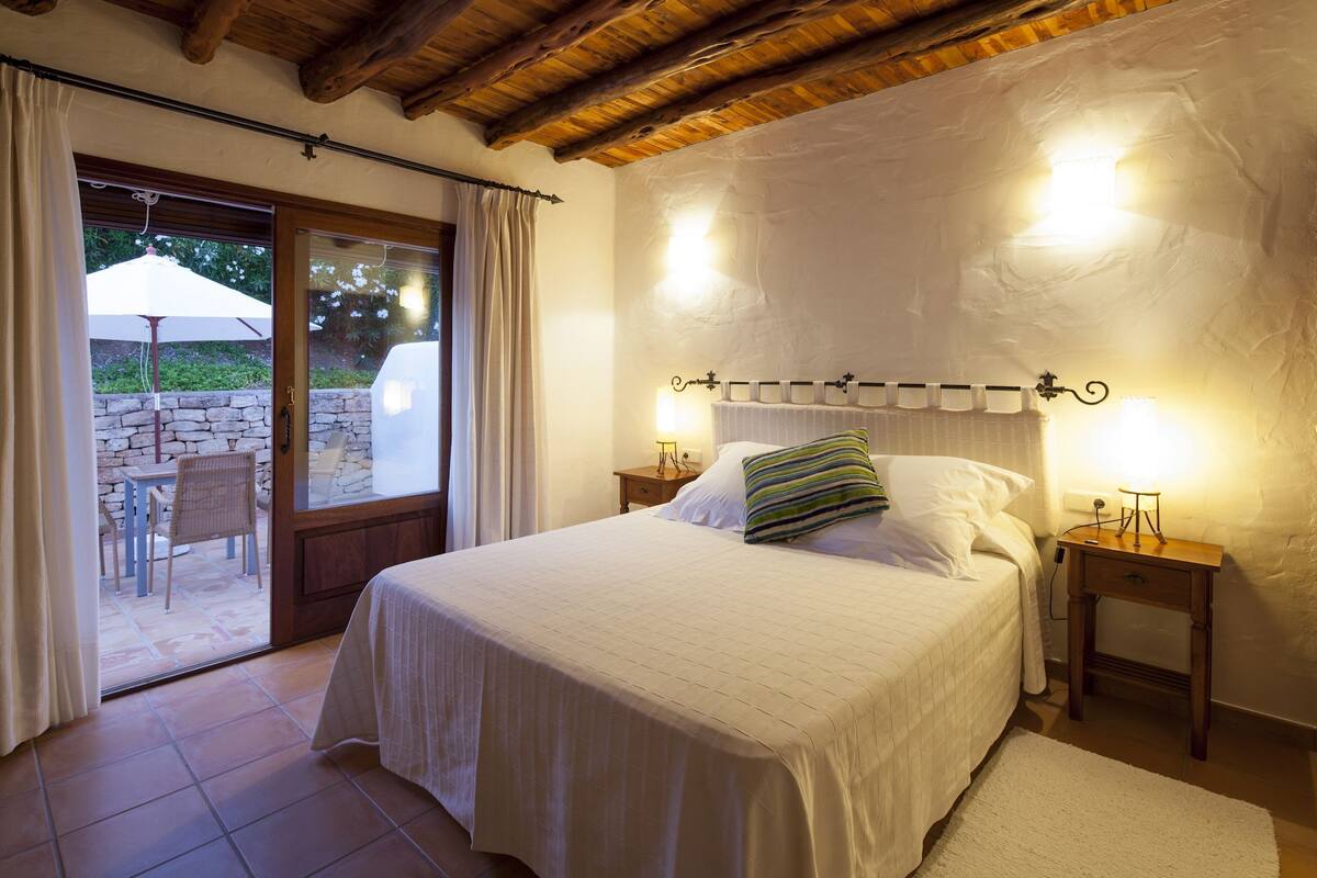 A bedroom with a bed, two bedside tables, a window with curtains, and a view of an outdoor patio.