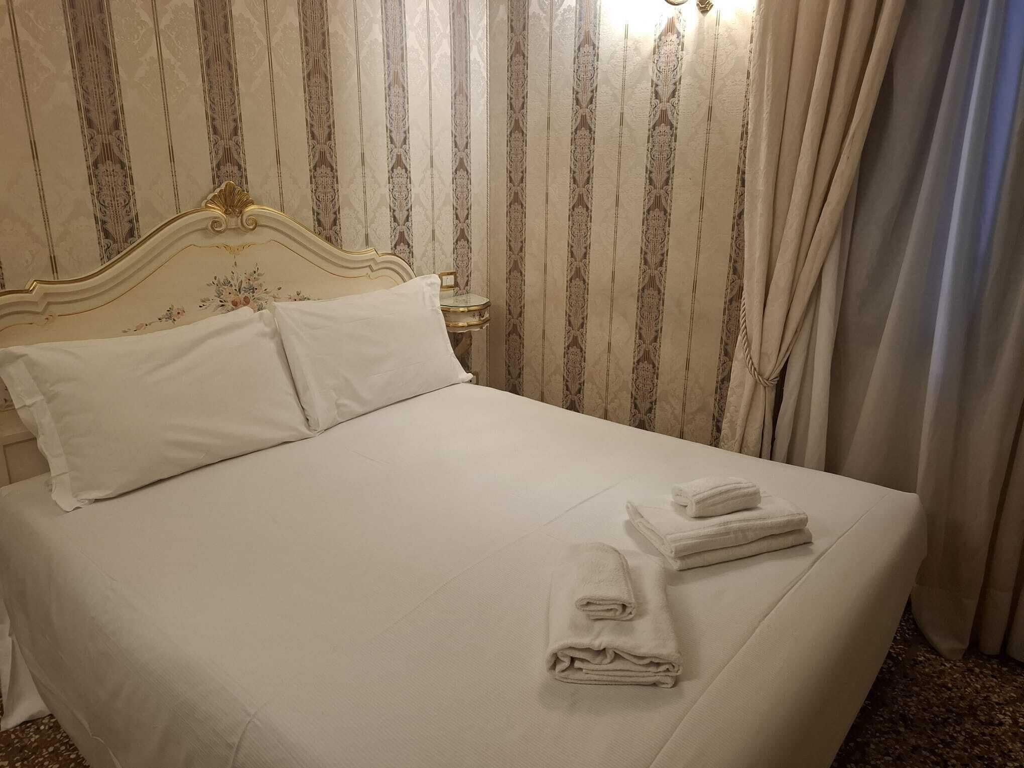 Una cama bien hecha con almohadas y toallas dobladas, una mesita de noche con lámpara, y una pared con rayas verticales.